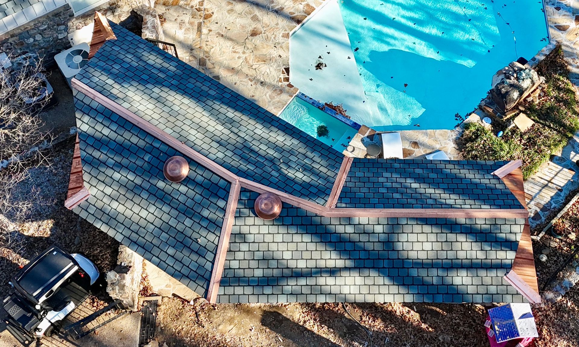 Aerial view of a dark shingled roof with copper-colored flashing, situated next to a bright blue swimming pool.