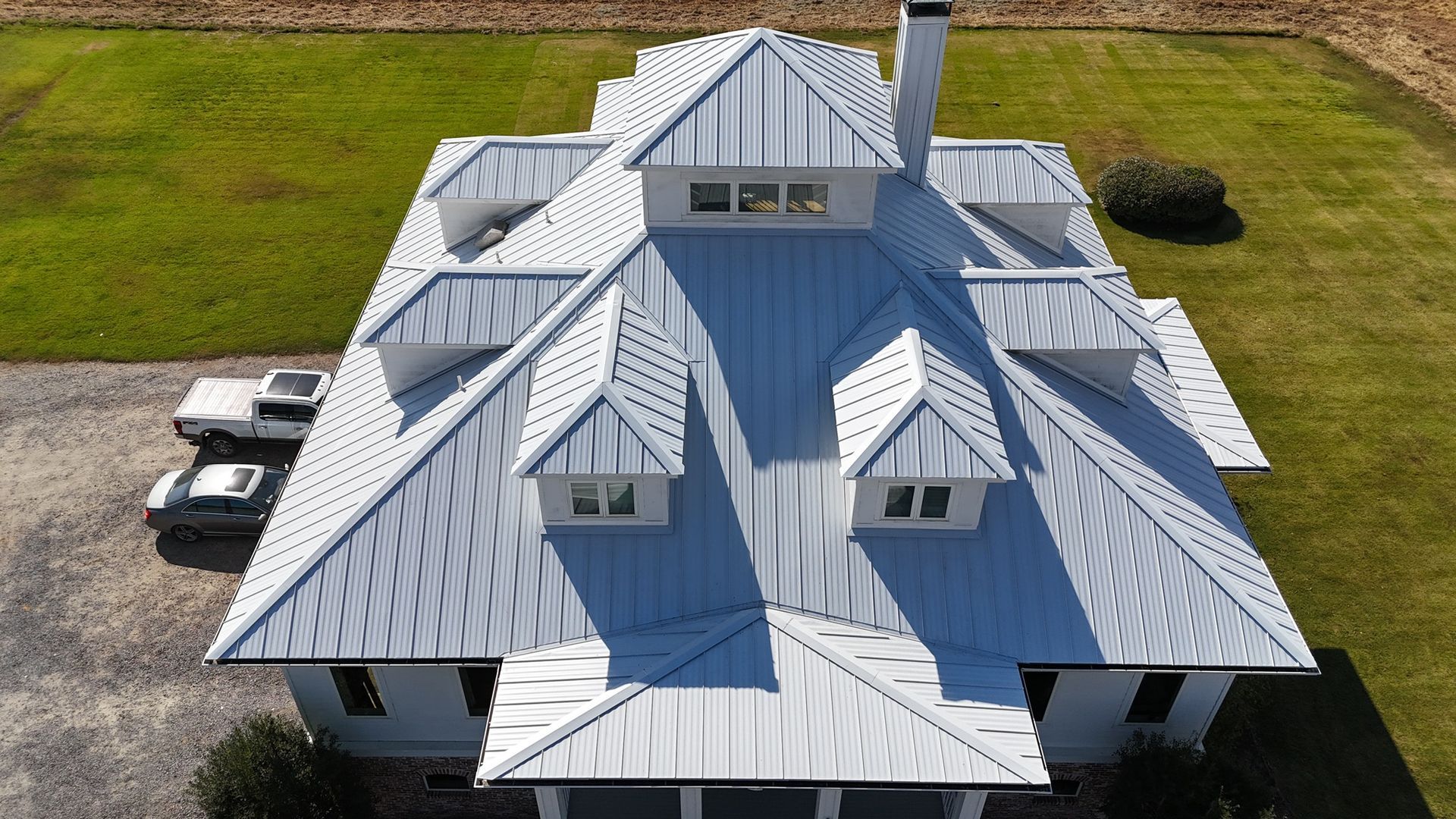 Aerial view of a white house with a silver metal roof, multiple gabled dormers, and a chimney, situated on a lawn.