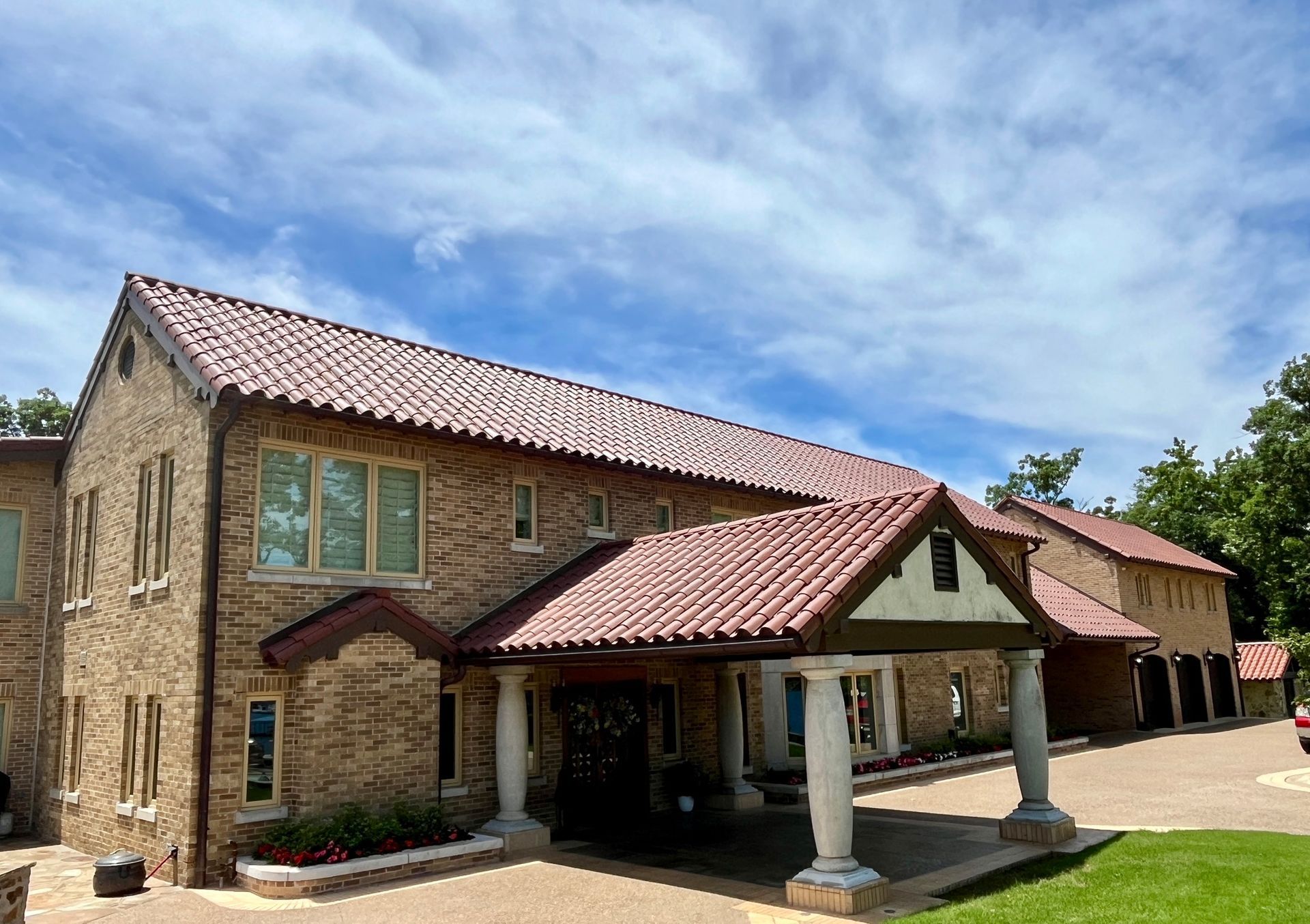 A two-story tan brick building with a brown tiled roof and a columned entrance portico under a bright, cloudy blue sky.
