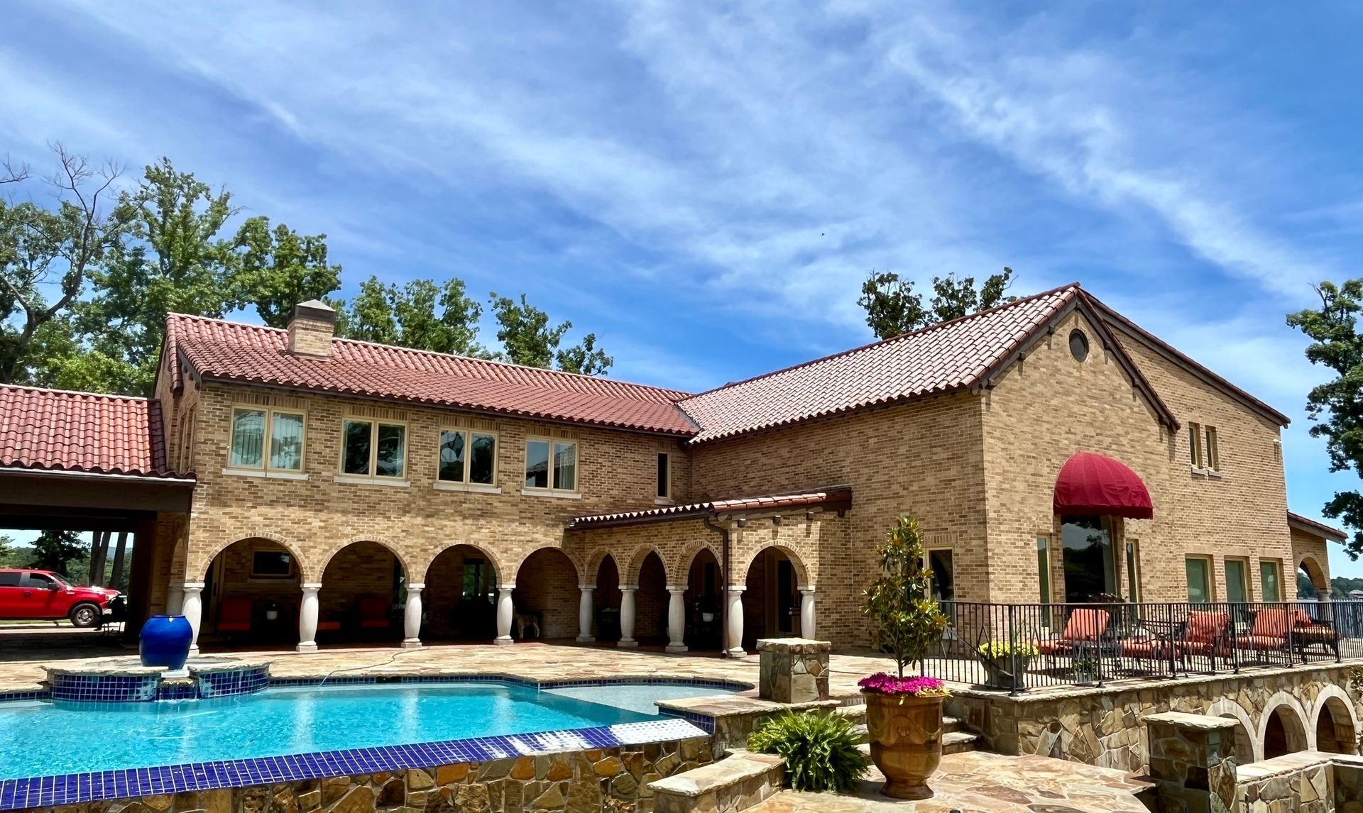 A tan stone villa with a red tile roof overlooks a blue swimming pool under a bright, sunny sky.