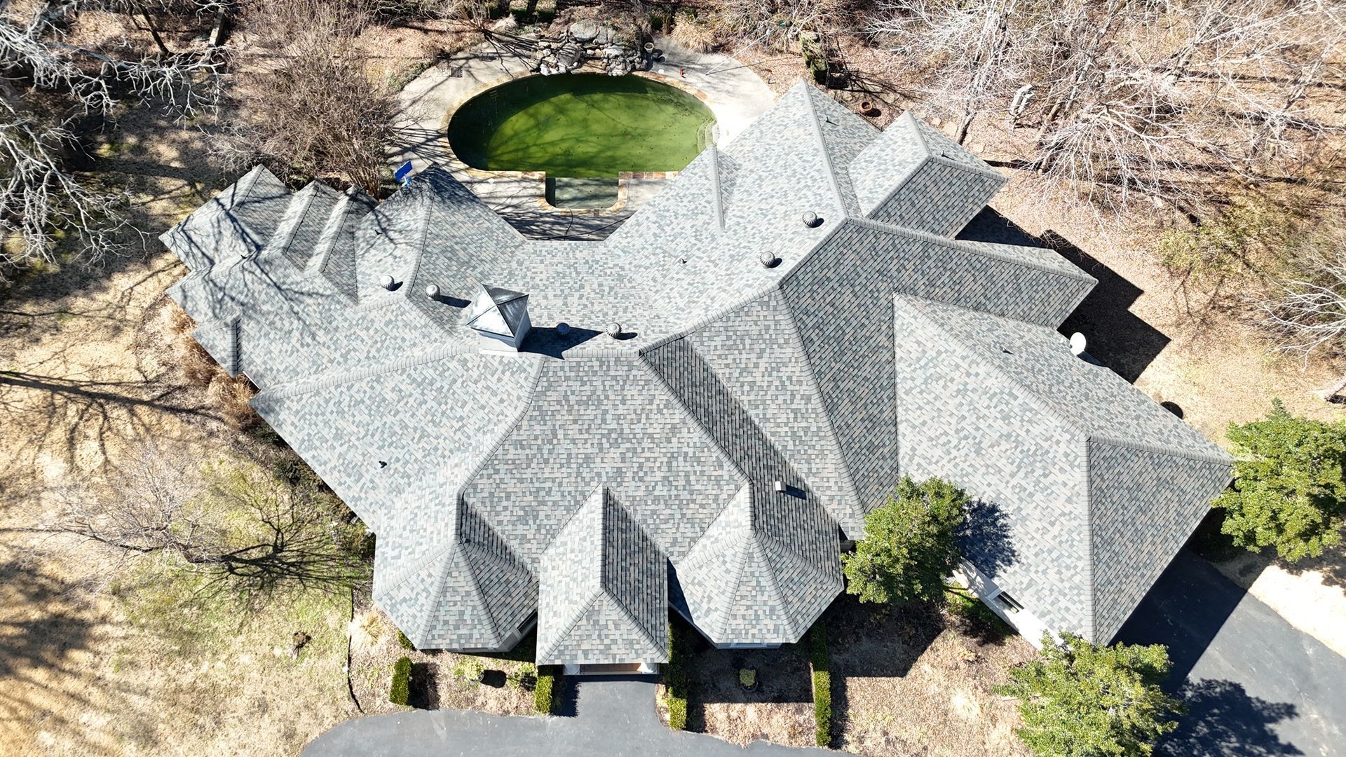 Aerial view of a sprawling house with a complex grey shingled roof, next to a circular green lawn in a wooded area.
