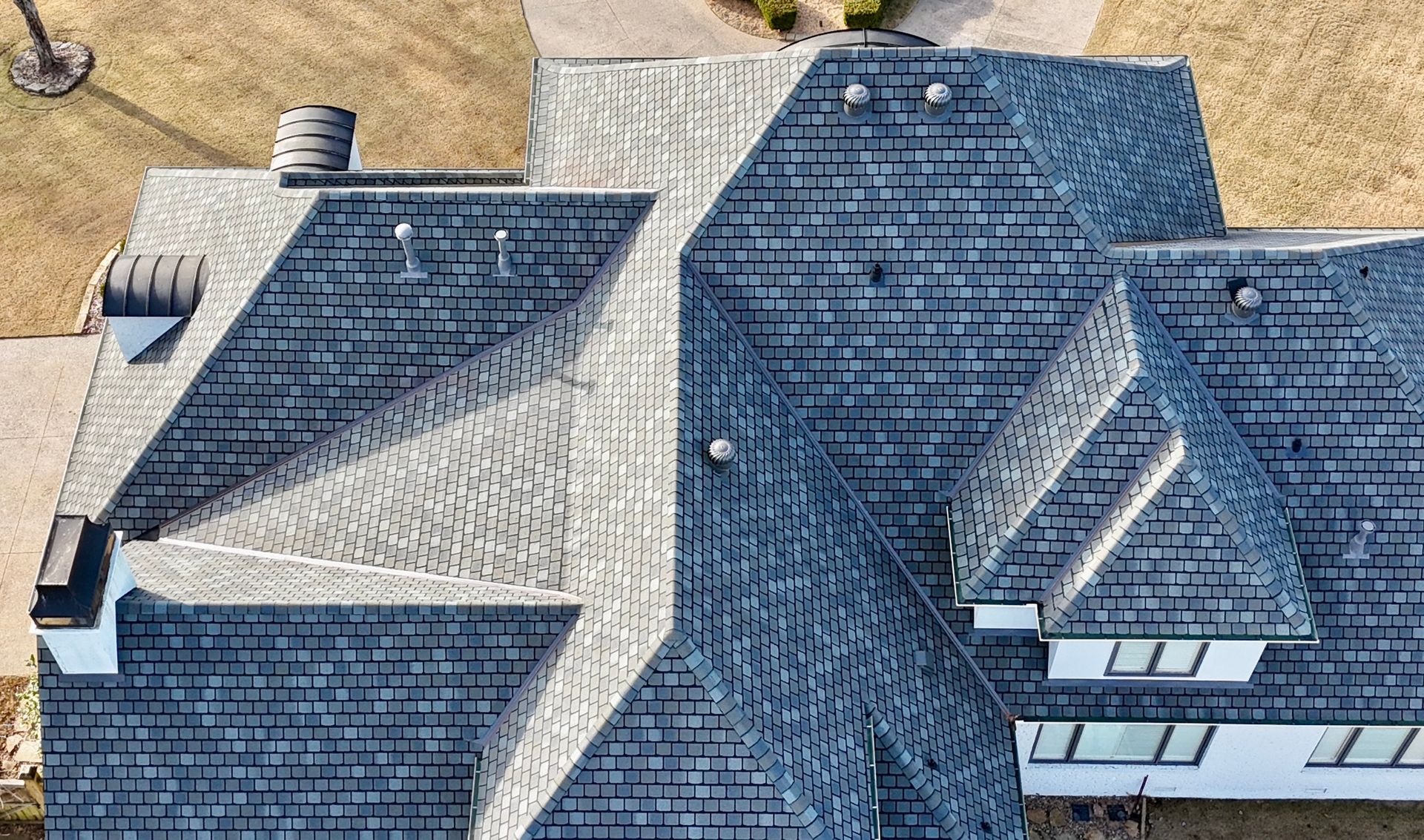 An aerial view of a dark shingled residential roof with varying sections, valleys, and skylights on a sunny day.