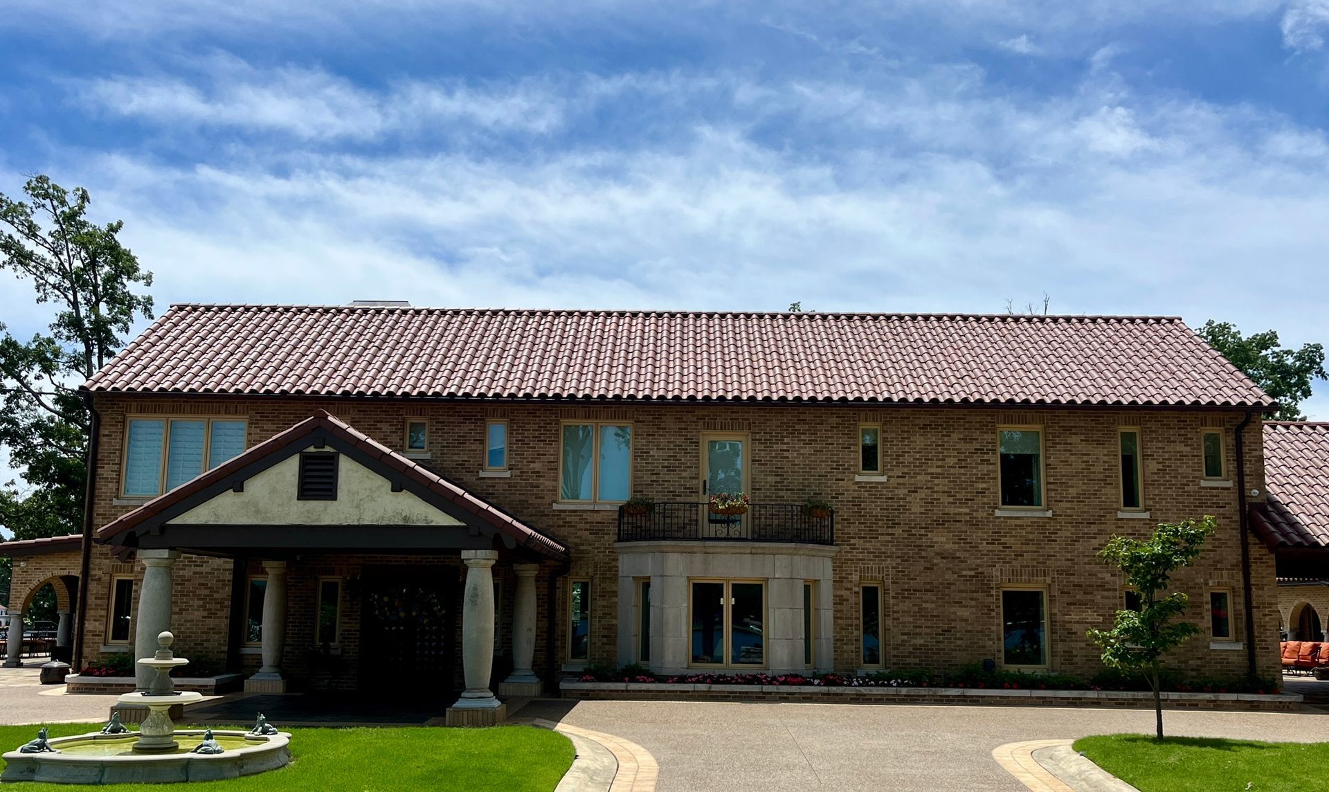 A large, two-story stone house with a tiled roof, a columned portico entrance, and a front fountain under a sunny sky.