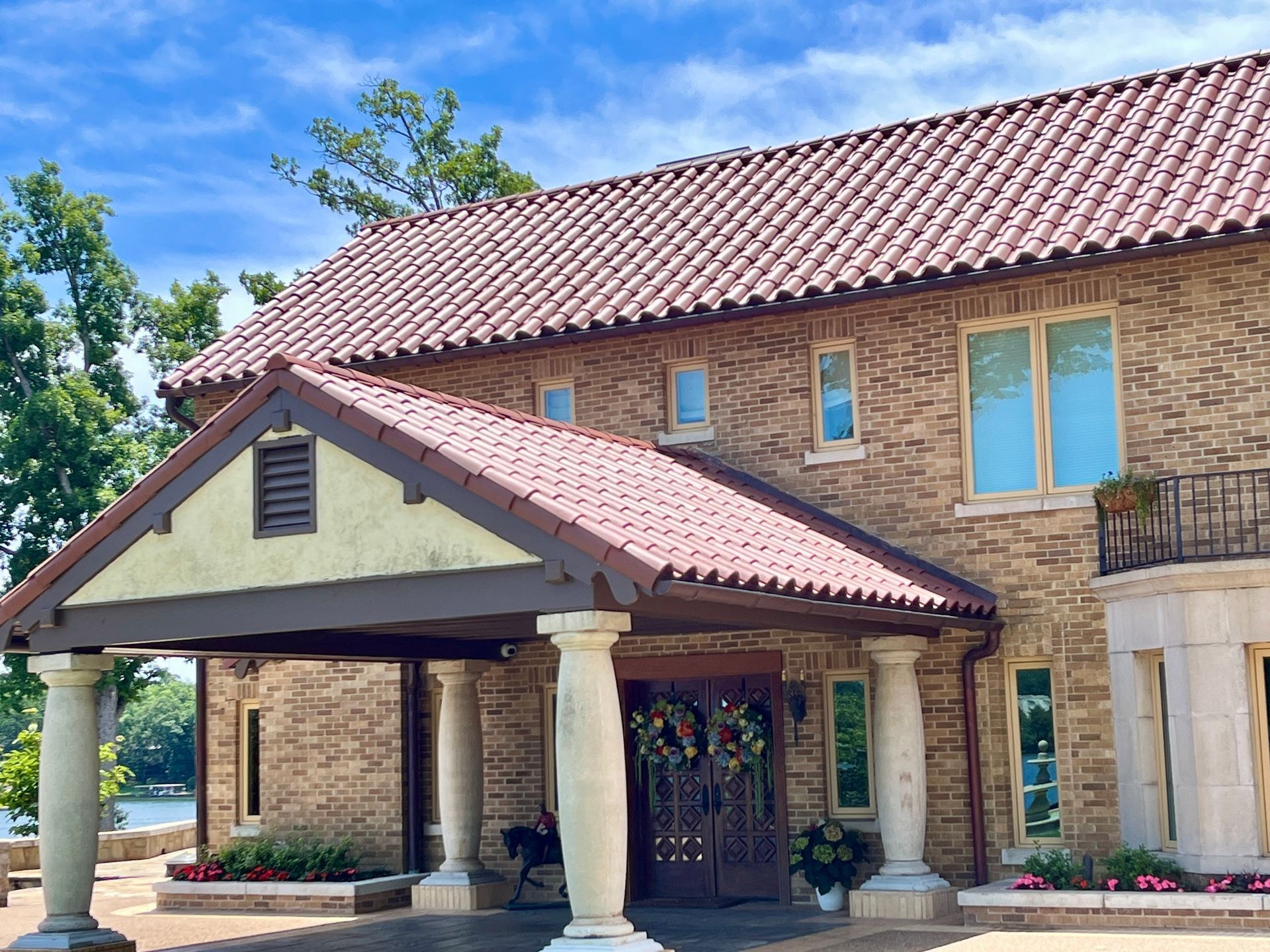 A large tan stone building with a red tile roof and a covered entryway supported by classical columns under a blue sky.