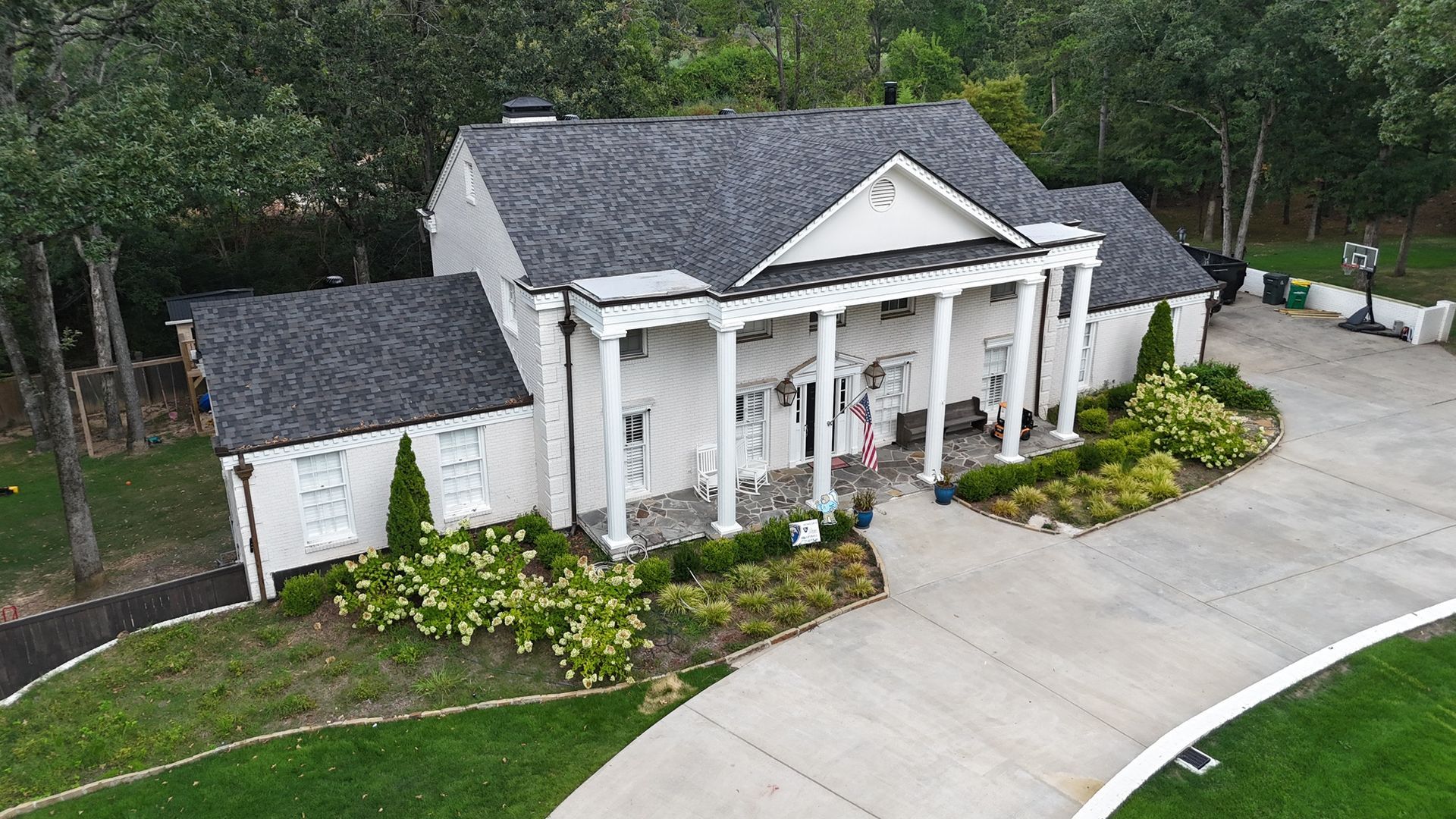 A white two-story house with a columned front porch, grey shingled roof, and a paved driveway, surrounded by trees.