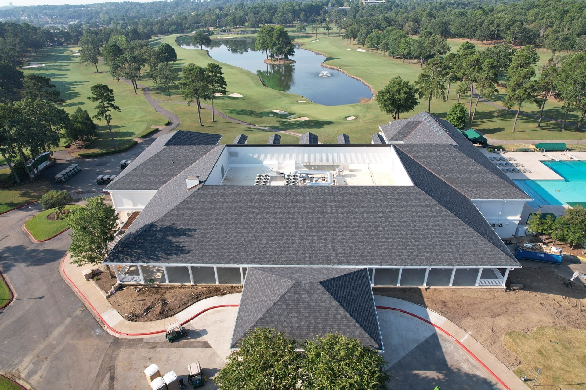 Aerial view of a large clubhouse with a gray shingled roof, next to a swimming pool and a golf course with a pond.