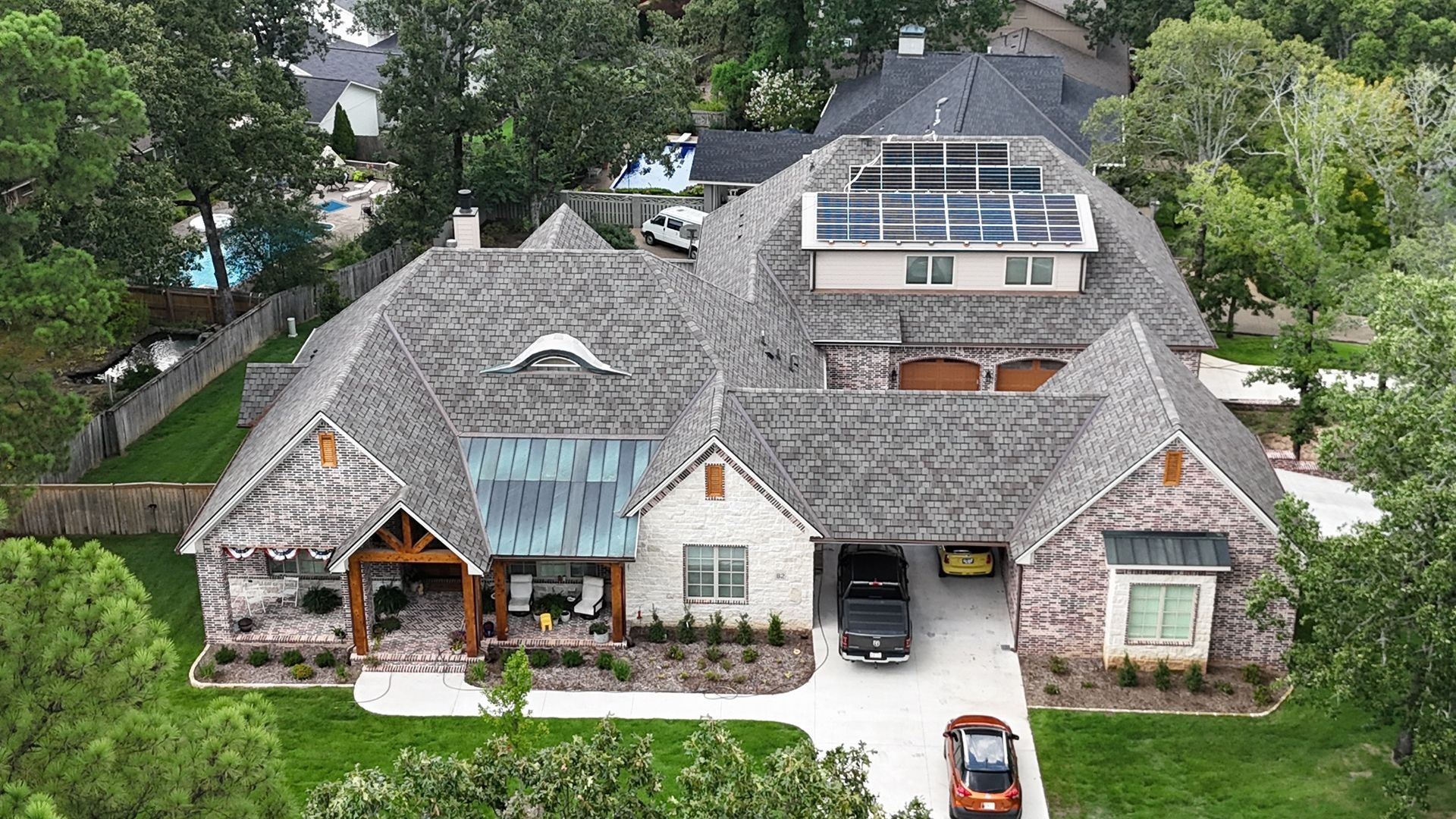 An aerial view of a large, gray-roofed brick house with a solar panel array and a driveway featuring two parked cars.