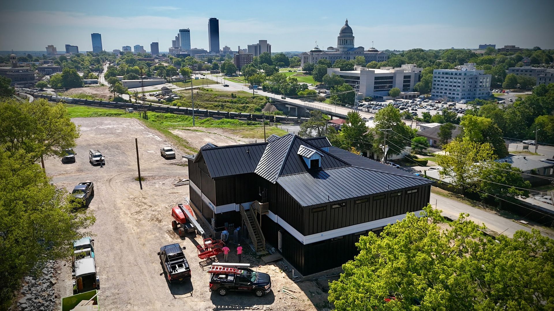Aerial view of a house under construction with black siding, parked vehicles, and a city skyline in the background.