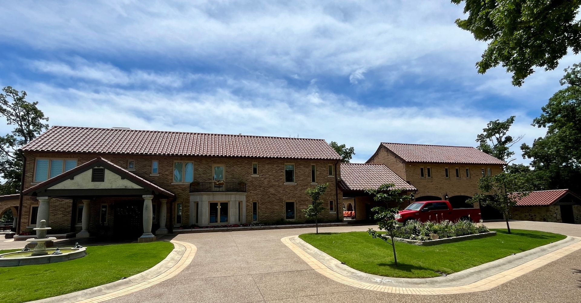 A large stone house with a red-tiled roof and a circular gravel driveway under a bright blue, cloudy sky.