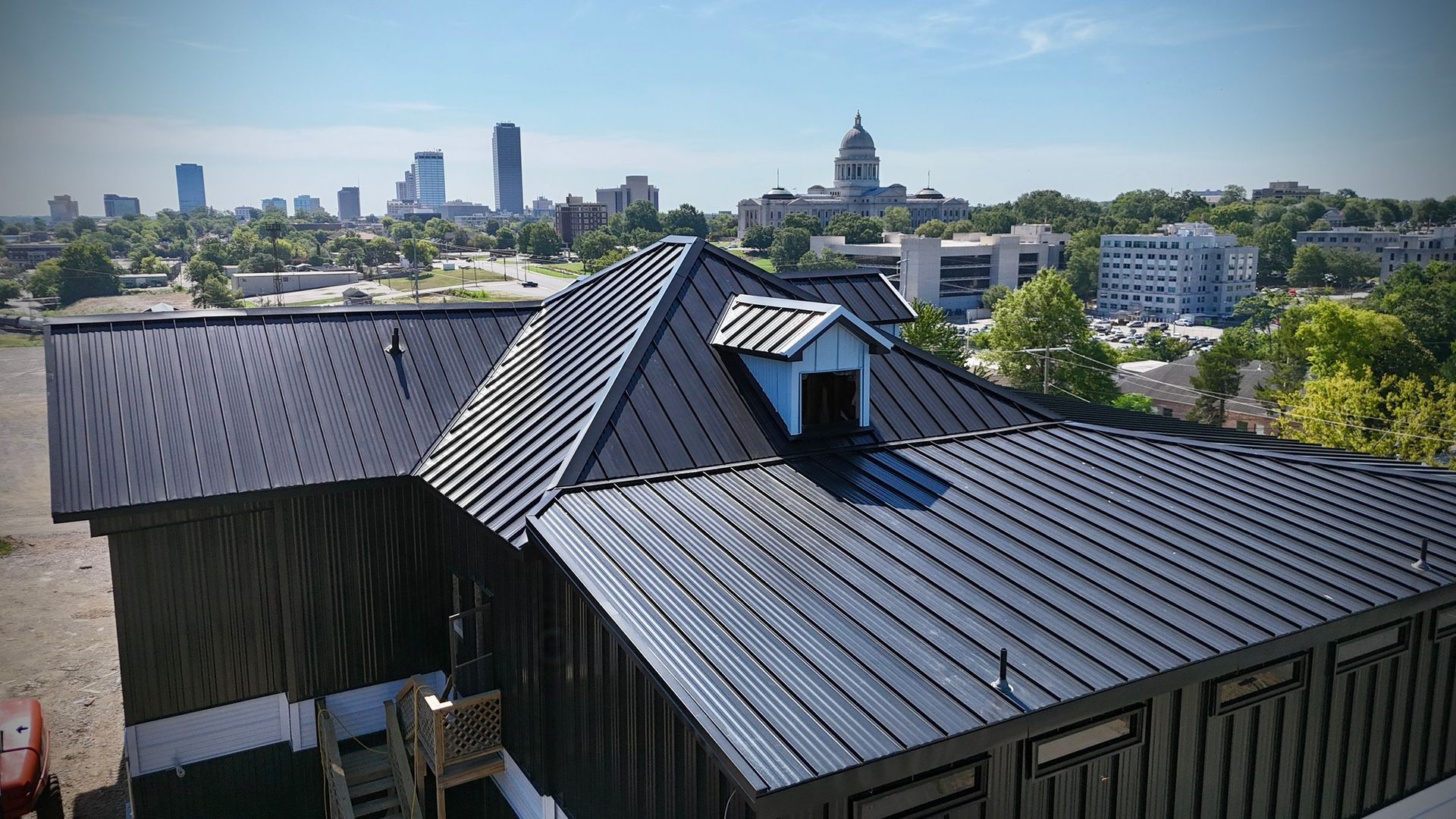 A high-angle view of a dark metal roof on a building, with a city skyline and a capitol dome visible in the distance.