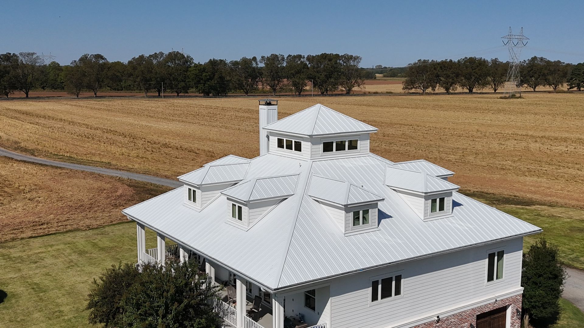Aerial view of a white, multi-roofed house in a rural field with a clear blue sky.